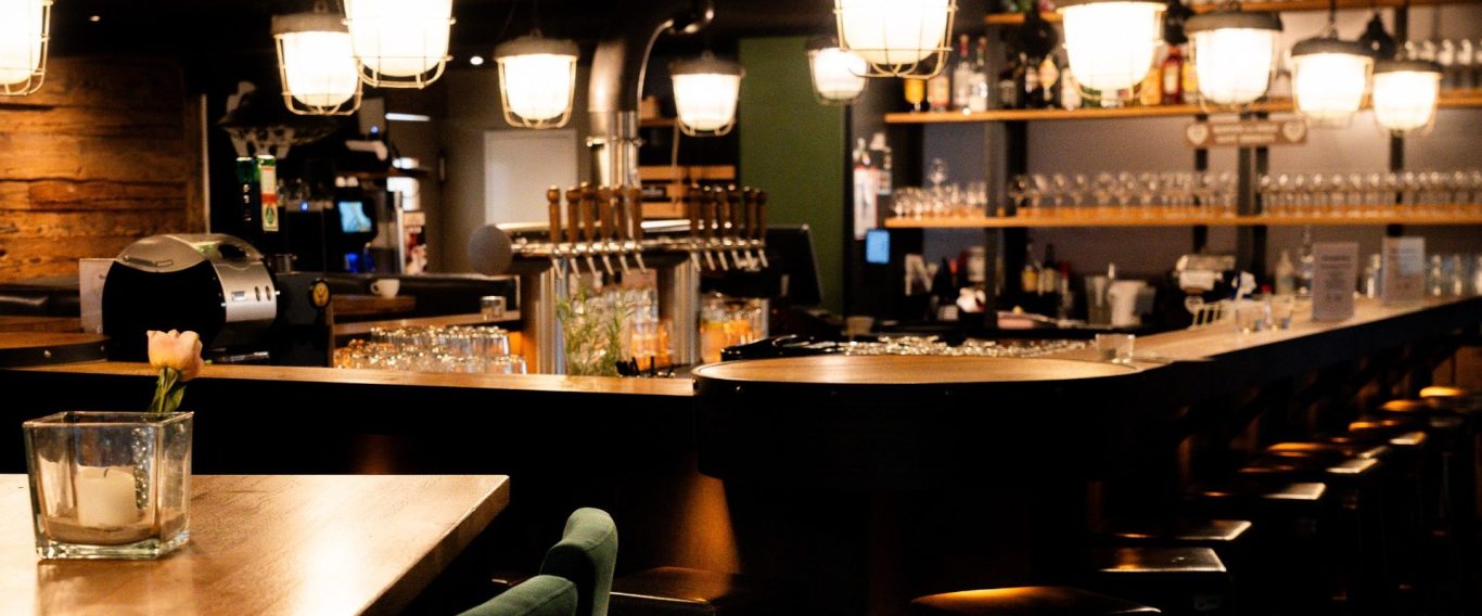 Moderner Tresen mit Holzdetails, Barhockern, beleuchtet von warmen Lampen und einem großen Gläserregal im Hintergrund.